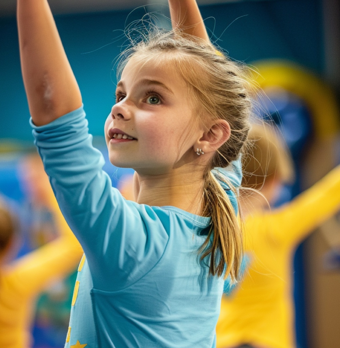 A young gymnast in a blue top reaching both arms overhead in a class