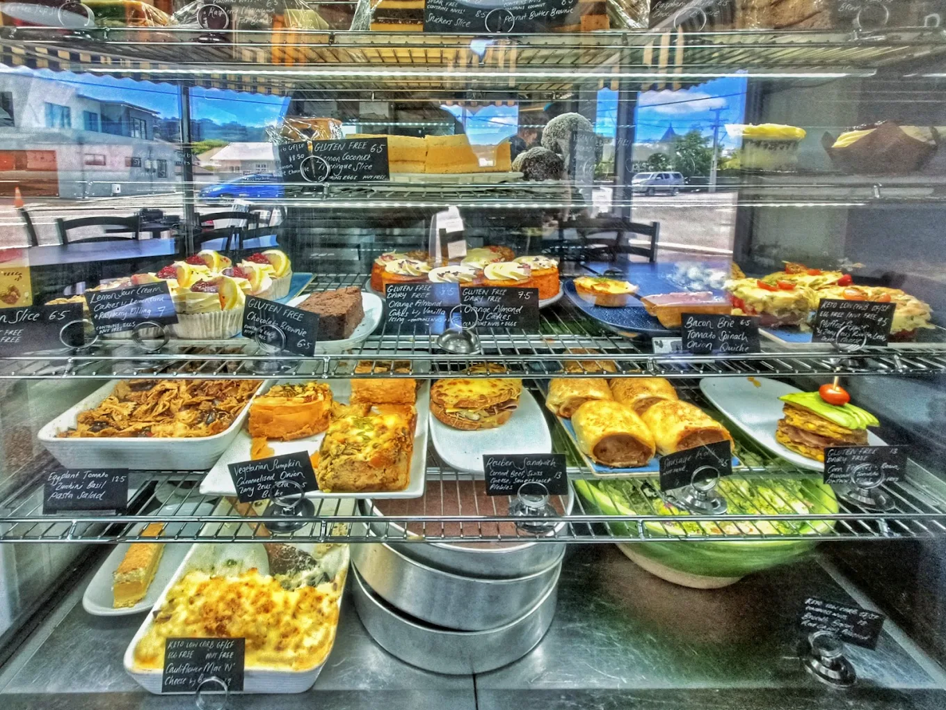 The cabinet full of slices, cakes, quiches, sausage rolls, salads and keto options, each with a chalk label.