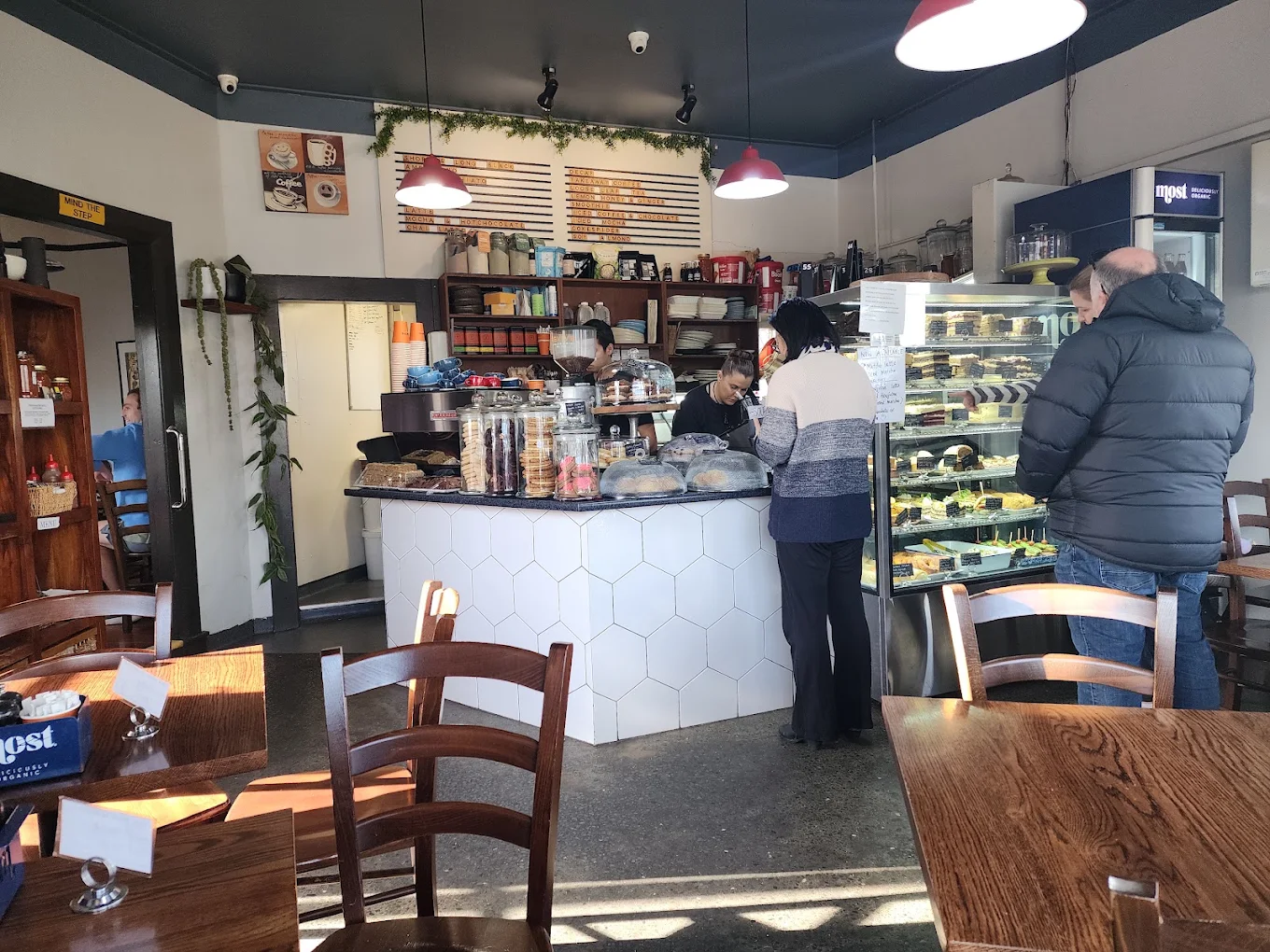 Inside the café: wooden tables, a white tiled counter, red pendant lights and the cabinet full of food.
