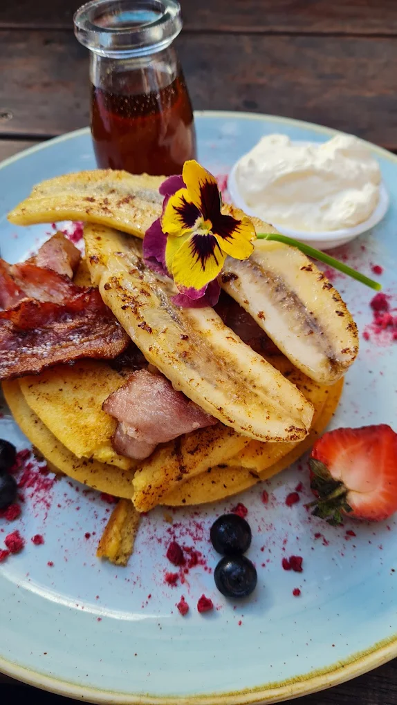 Pancakes topped with grilled banana, bacon, berries and an edible flower, with maple syrup and cream.